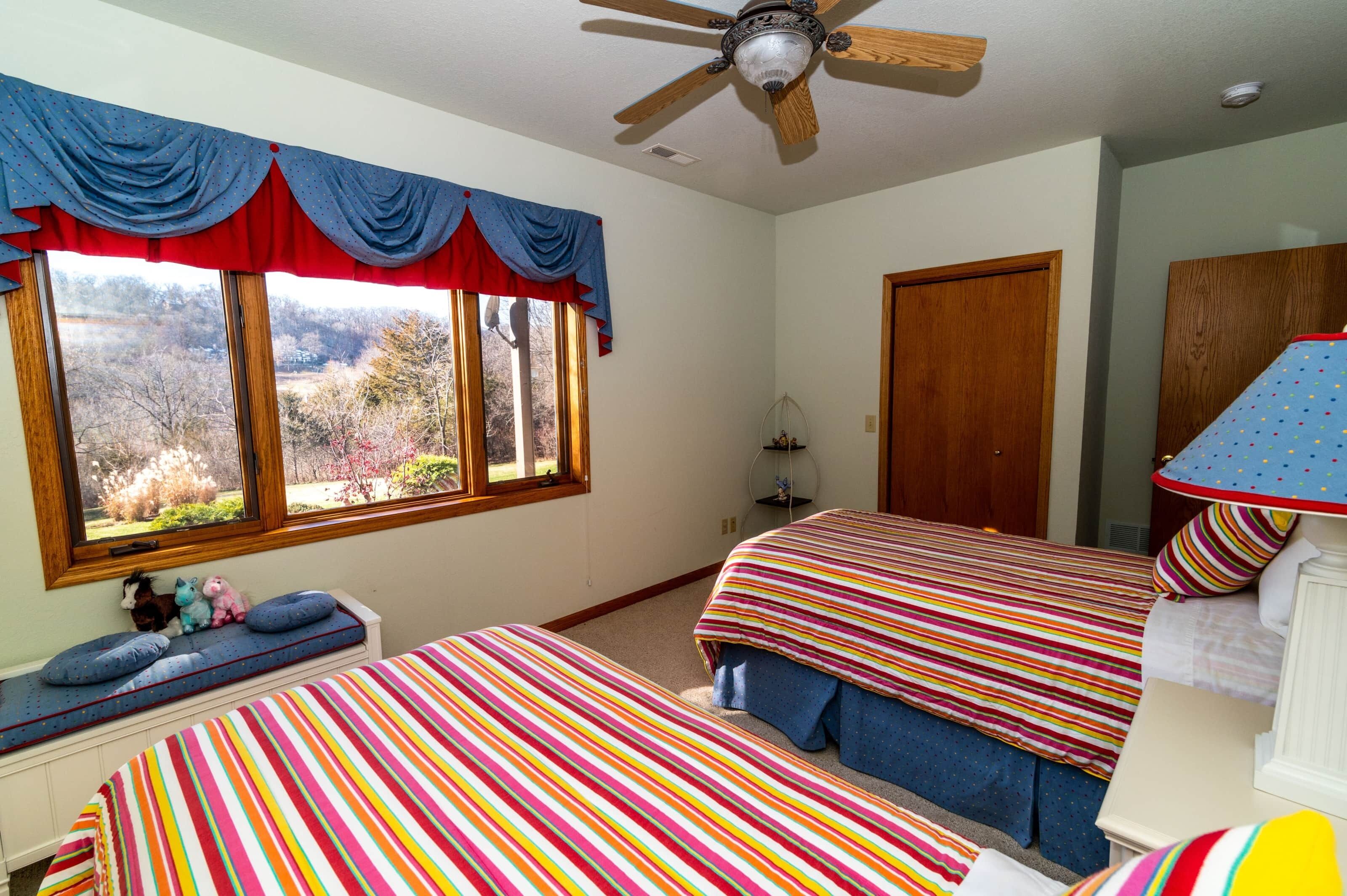 Une chambre avec deux lits, un ventilateur de plafond, une fenêtre avec des rideaux et une vue sur l’extérieur.