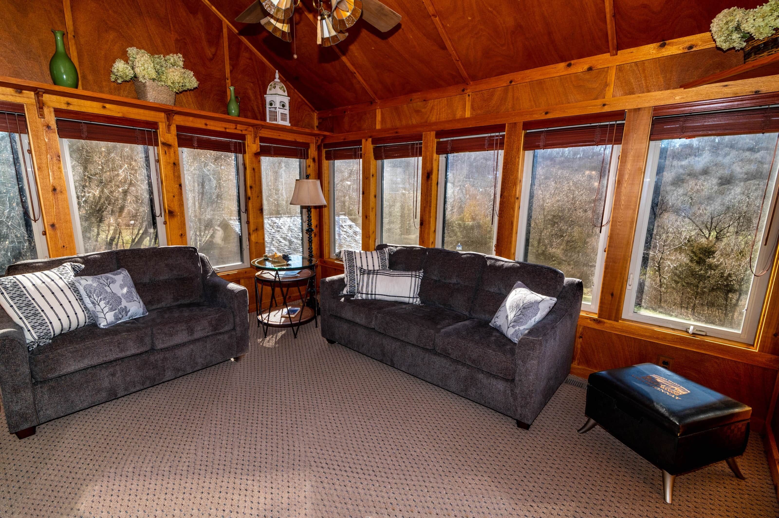 A cozy room with two dark sofas, a small round table, and a lamp. Large windows offer a view of trees outside.
