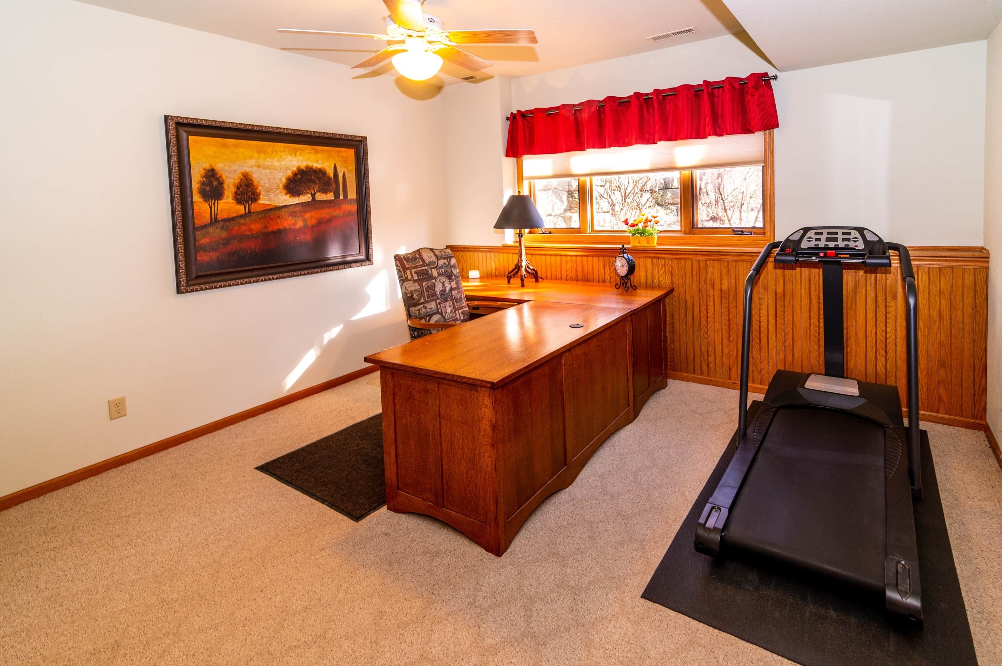 A home office with a desk, chair, treadmill, and a painting on the wall.