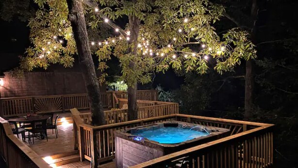 A hot tub surrounded by a wooden deck with string lights, a wooden fence, and outdoor furniture. - The Owl Barn - Most Unique Cabin in The Smokies. Hot Tub, Family & Pet Friendly (Sevierville)