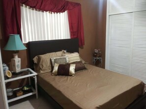A bedroom with a bed, a nightstand, a lamp, a window with curtains, and a closed door. - Bon Soleil (Miramar)