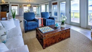 Un salon spacieux avec un canapé blanc, deux fauteuils bleus, une table basse en bois et une vue sur l’océan grâce à de grandes fenêtres.