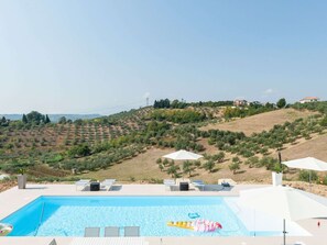 Ein Schwimmbecken mit Liegestühlen und Sonnenschirmen, umgeben von Olivenhainen und einer hügeligen Landschaft.