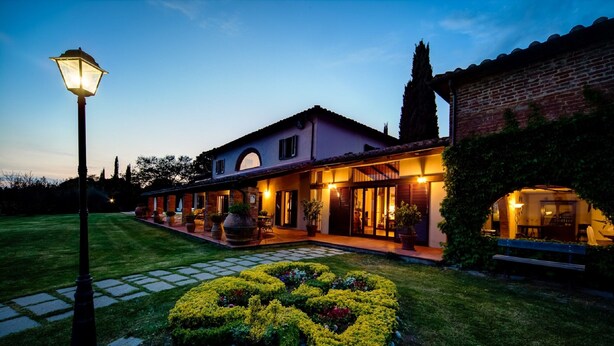 A well-lit, two-story house with a paved entrance, a landscaped garden with a circular flower bed, and a brick wall on the right. - Villa by Lake Trasimeno With Scenic Views (Castiglione del Lago)