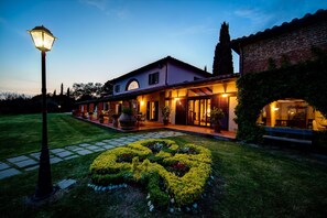 A well-lit, two-story house with a paved entrance, a landscaped garden with a circular flower bed, and a brick wall on the right. - Villa by Lake Trasimeno With Scenic Views (Castiglione del Lago)