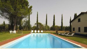 A rectangular swimming pool with a brick border, surrounded by a grassy area with trees and a white building with windows. - Villa by Lake Trasimeno With Scenic Views (Castiglione del Lago)