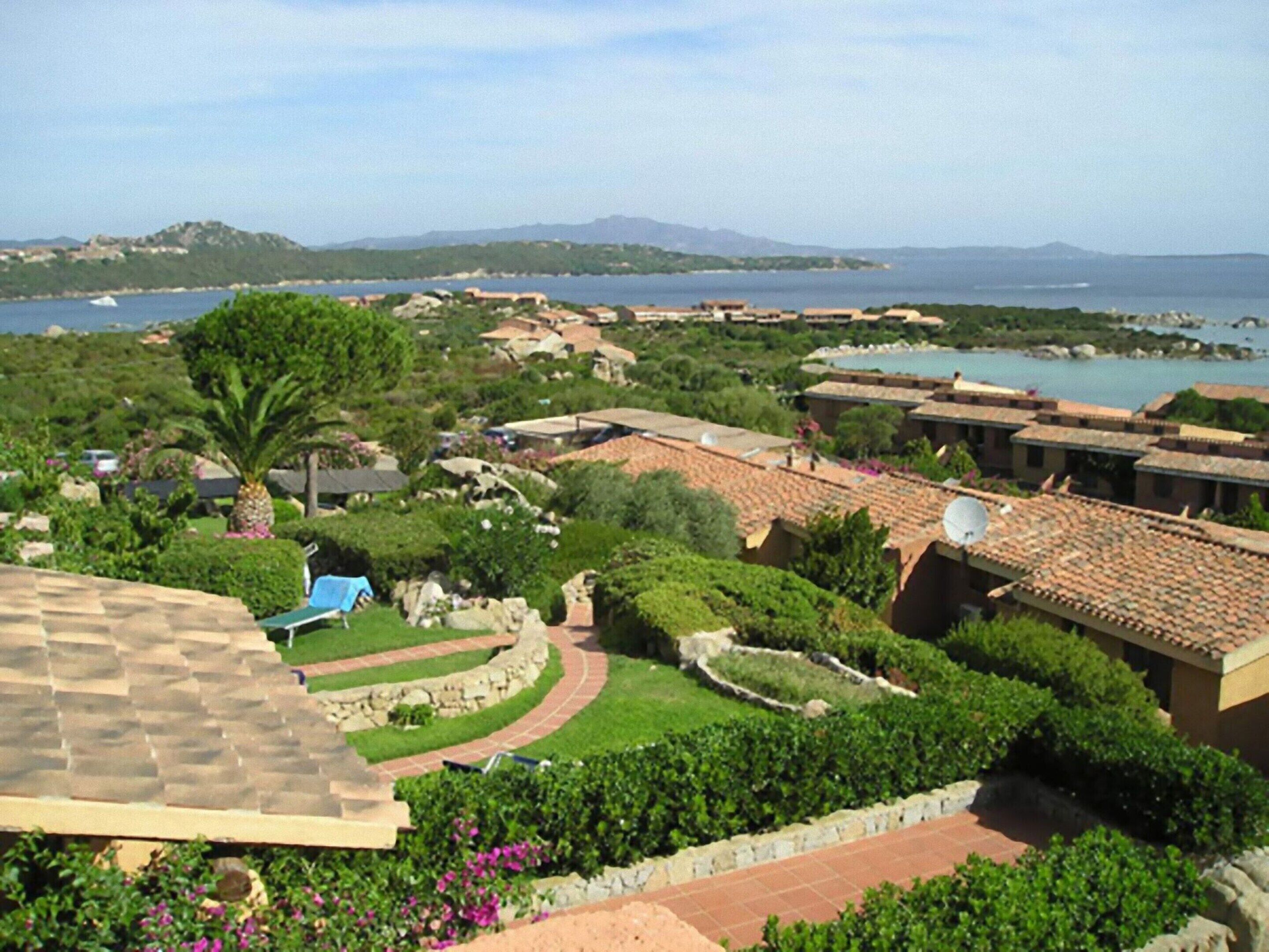 A coastal resort with terracotta-roofed buildings, lush greenery, and a view of the sea and distant mountains.