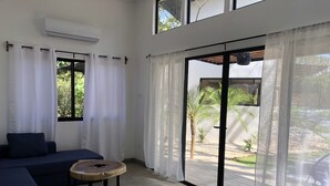 A modern living room with a sofa, a small round table, and a sliding glass door leading to an outdoor area with plants. - Honeymoon apt with mini fridge and pool view (Playa junquillal)