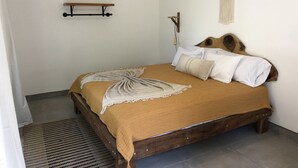 A bedroom with a wooden bed, white pillows, a beige blanket, a wooden headboard, a wall-mounted air conditioner, and a small shelf. - Honeymoon apt with mini fridge and pool view (Playa junquillal)