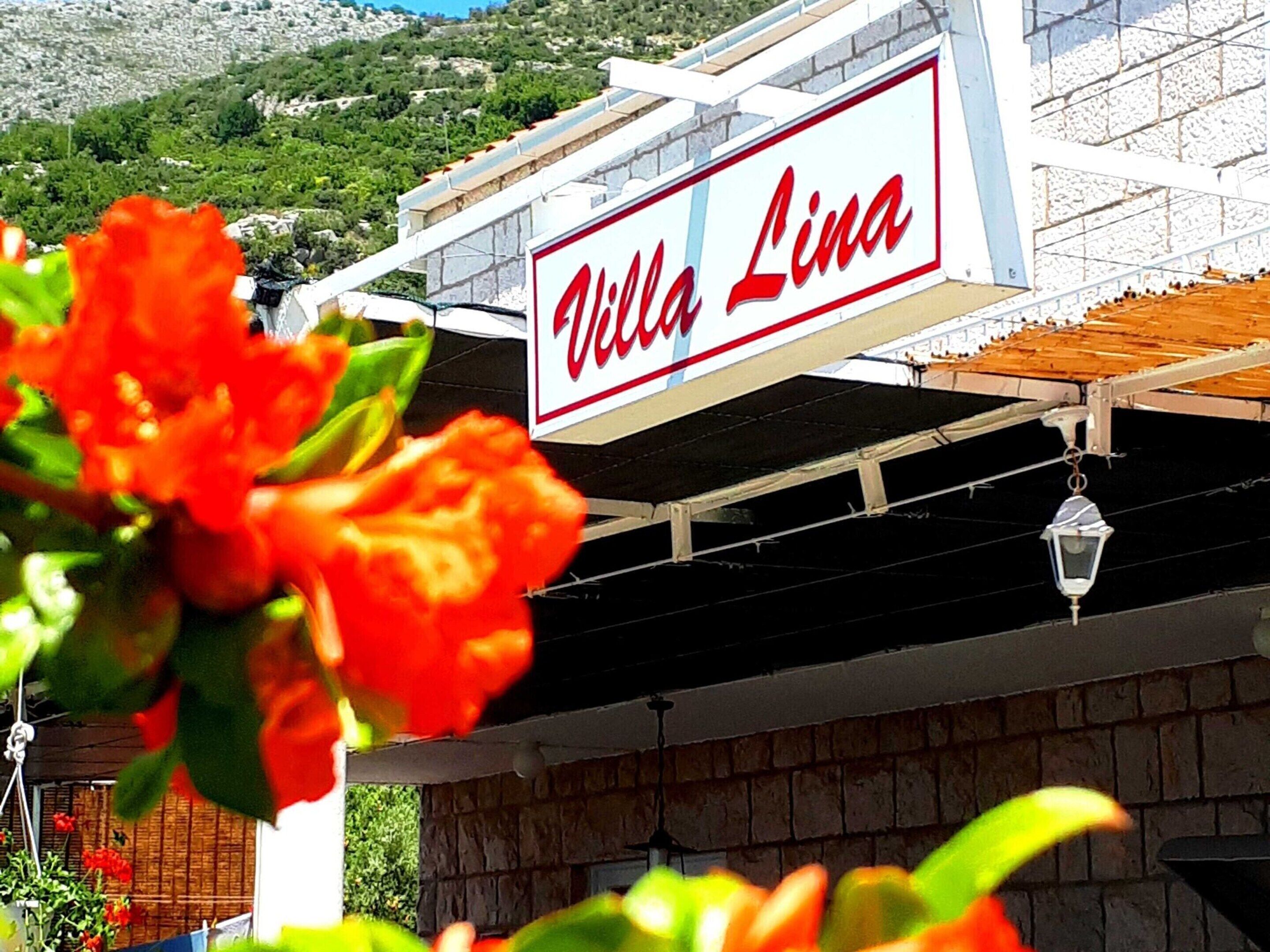 A sign for "Villa Lina" with a view of a building and greenery in the background.