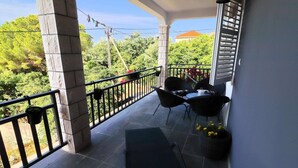 A covered outdoor area with a table and chairs, a potted plant, and a view of greenery and buildings in the background. - Apartment Marjana Prizba - Two Bedroom Apartment With Terrace and Partial Sea View (Blato)