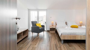 A modern bedroom with a wooden floor, a grey armchair with a yellow pillow, a bed with white linens, and two bedside lamps. - Fancy Apartments Dream - Comfort One Bedroom Apartment With Balcony and Sea View (Smokvica)