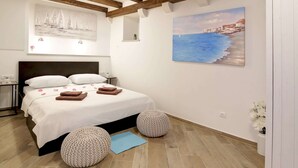 A bedroom with a bed, wooden beams, a painting of sailboats, a painting of a coastal scene, and two round ottomans. - Apartment City Gem - Studio (Dubrovnik)