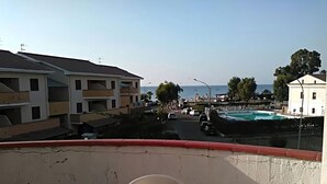 A balcony with a round table and two chairs, overlooking a street with buildings and a pool. - Holiday Home Near Marina di Mandatoriccio Beach (Mandatoriccio)