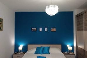 Un dormitorio con una cama grande, dos mesitas de noche, una araña y cuatro cuadros en la pared.