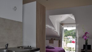 Eine moderne Küche mit Spüle und Herd, ein Bett mit violetter Bettwäsche und Blick auf einen Balkon mit einer roten Blume.