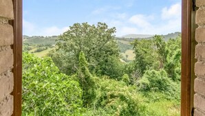A view through a window with a brick wall and wooden beams, overlooking a lush green landscape with trees and rolling hills. - Belvilla by OYO Bilocale Monolocale 2adeg Piano (Assisi)