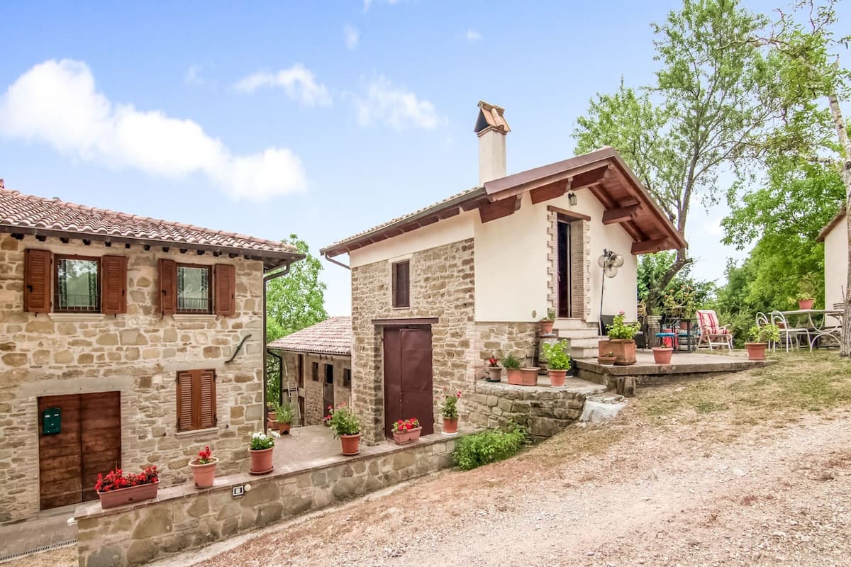 Un bâtiment rustique en pierre, doté d’un toit en tuiles, d’un petit porche orné de plantes en pot et d’une allée gravillonnée.