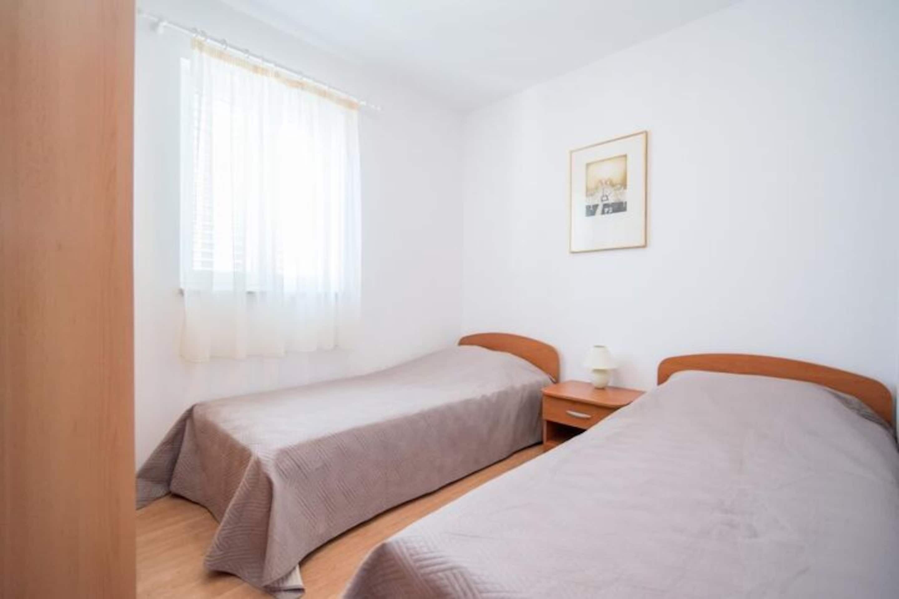 A small, clean room with two single beds, a nightstand, a framed picture on the wall, and a window with sheer curtains.
