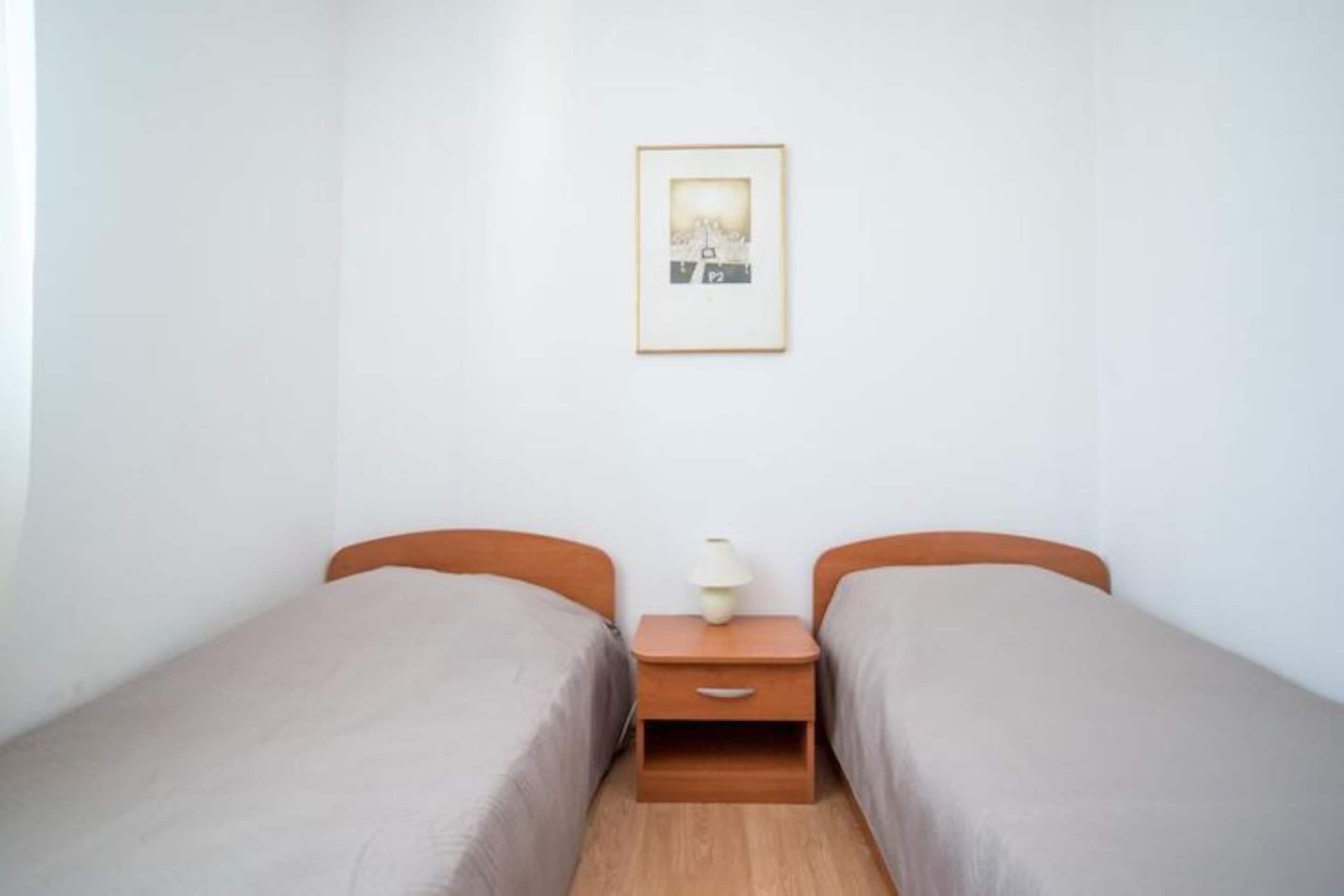 Ein kleines, sauberes Zimmer mit zwei Einzelbetten, einem Nachttisch mit Lampe und einem gerahmten Bild an der Wand.
