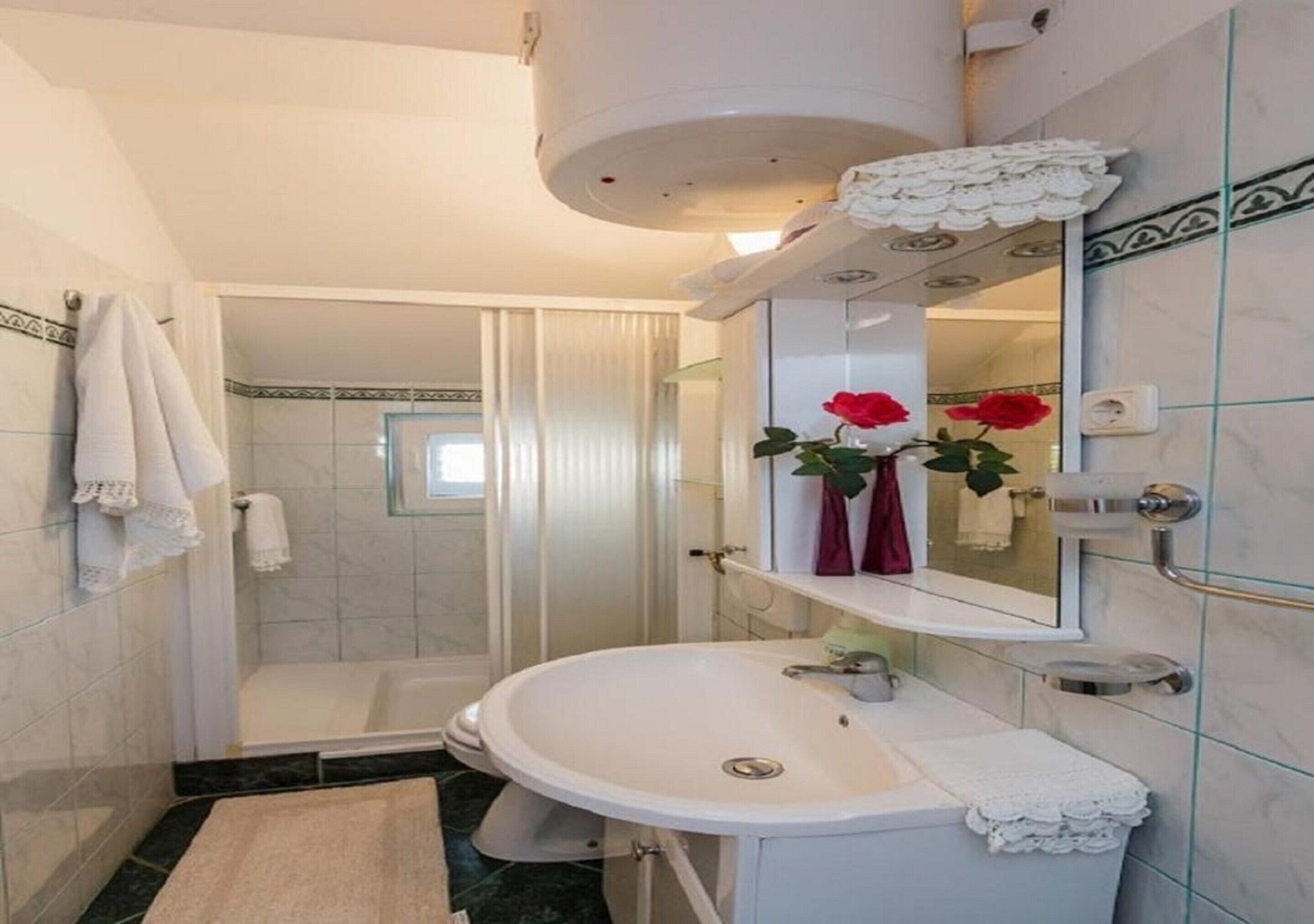 Un baño con lavamanos blanco, cabina de ducha de vidrio y espejo con un florero de rosa roja.