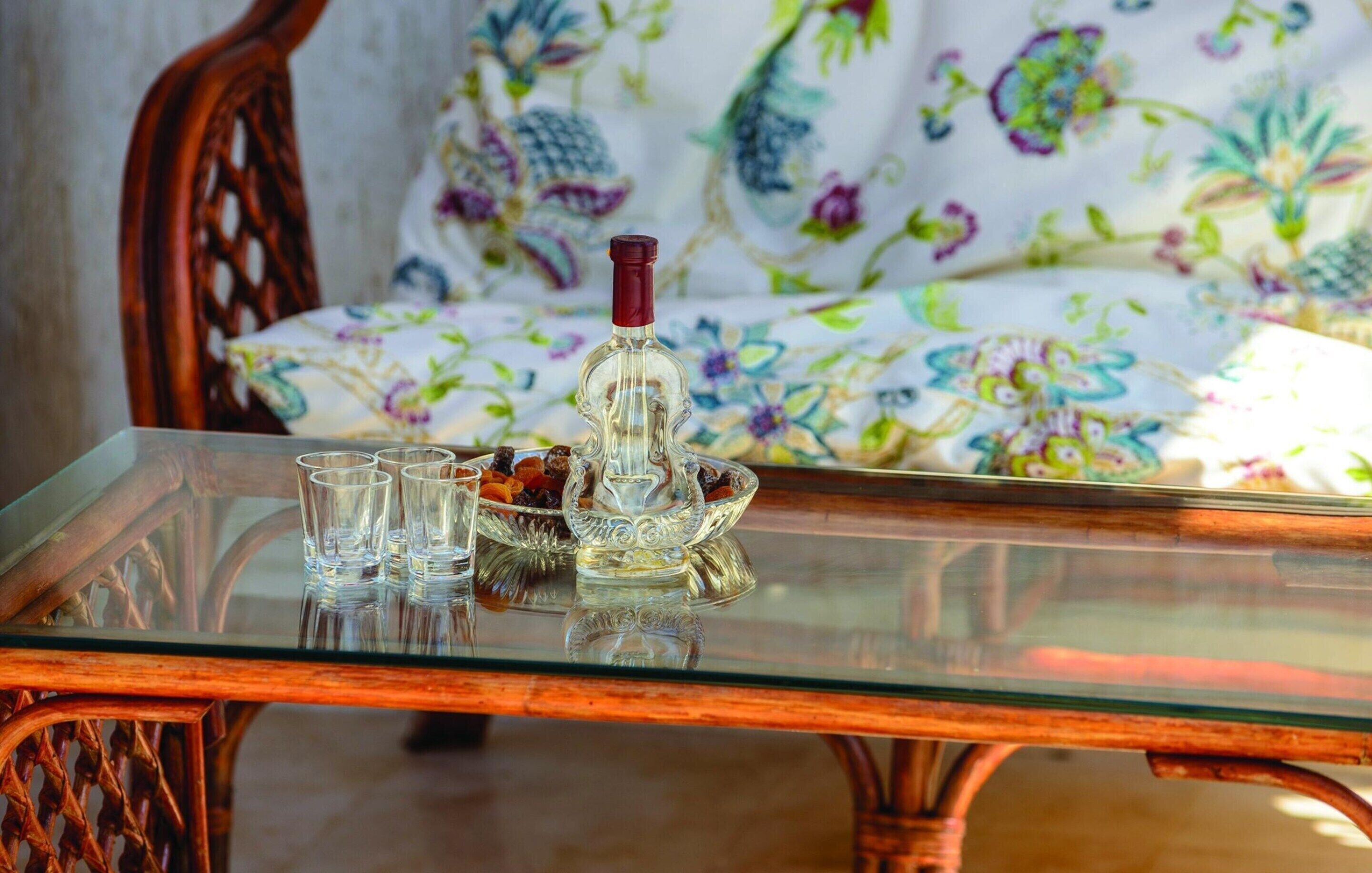 A glass-top table with a bottle and shot glasses, a bowl of fruit, and a floral-patterned sofa in the background.