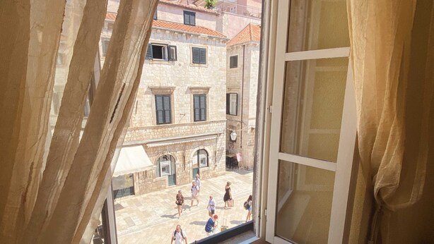 A window with a view of a courtyard with stone buildings, a few people walking, and a table with a white umbrella.
