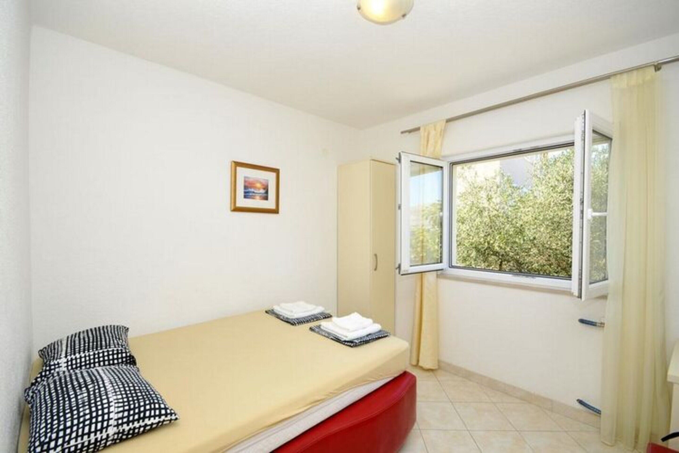 A small, single-bed room with a window, a framed picture on the wall, and a red bench.