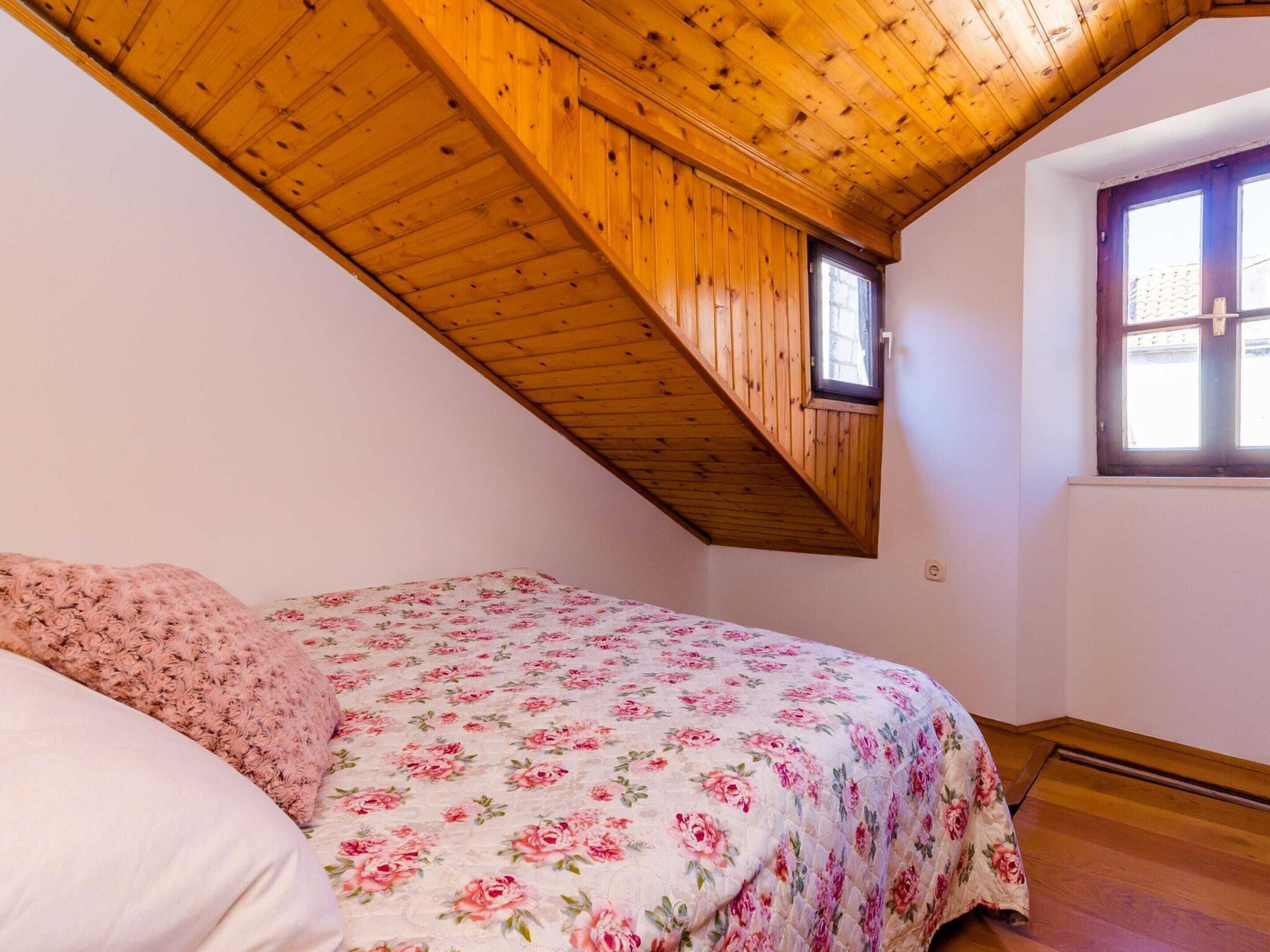 A bedroom with a sloped wooden ceiling, a bed with a floral bedspread, and a window with blinds.