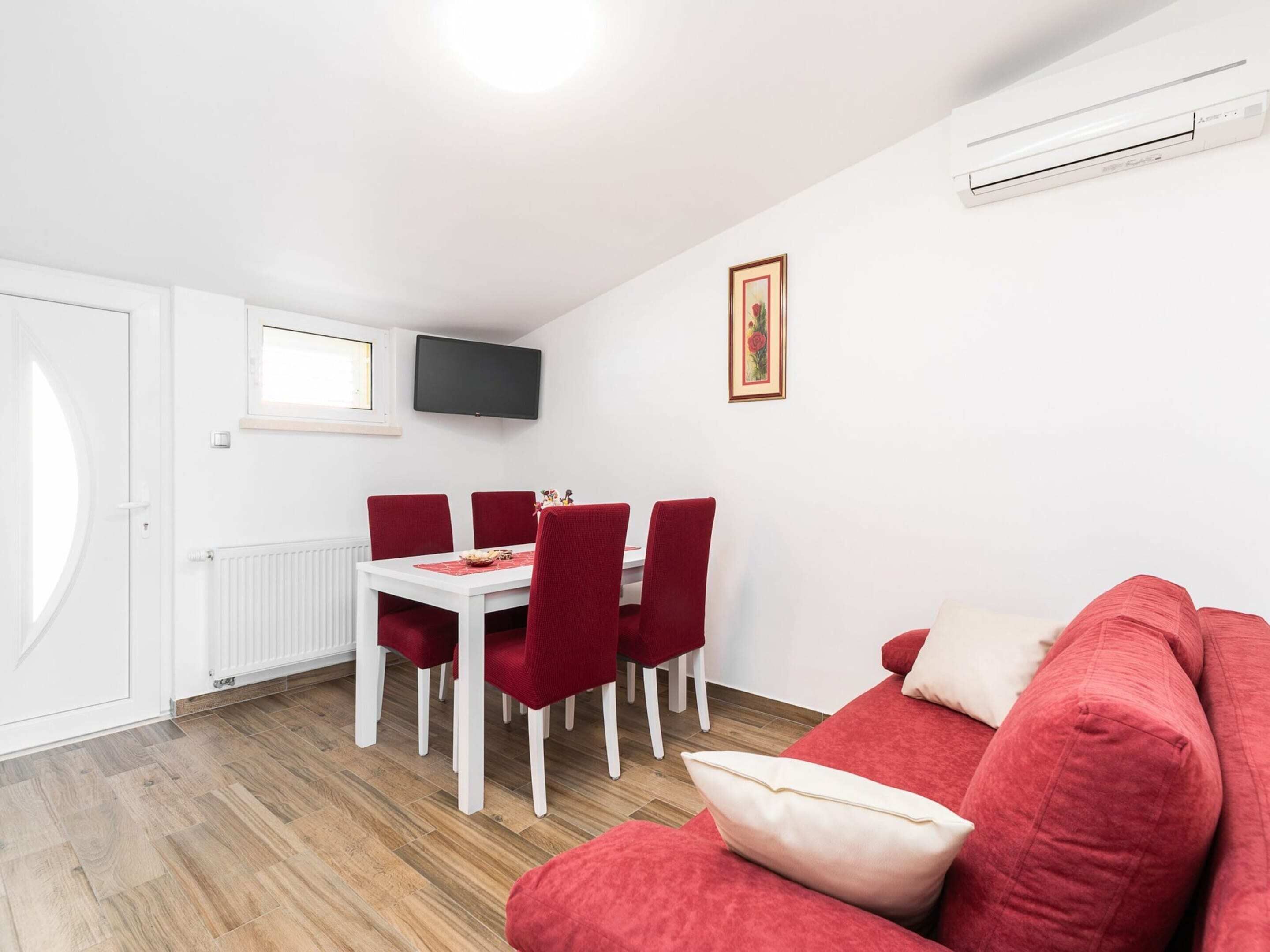 A modern living room with a red sofa, a dining table with red chairs, a flat-screen TV mounted on the wall, and a radiator under the window.