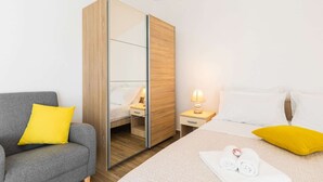 Ein Hotelzimmer mit grauem Sofa, einem Bett mit weißen und gelben Kissen, einem Holzschrank und einem Nachttisch mit Lampe.