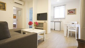A living room with a sofa, a coffee table with flowers, a TV, and a kitchen area with a fridge and a small table. - Apartment Goge - One-bedroom Apartment With Balcony (Dubrovnik)