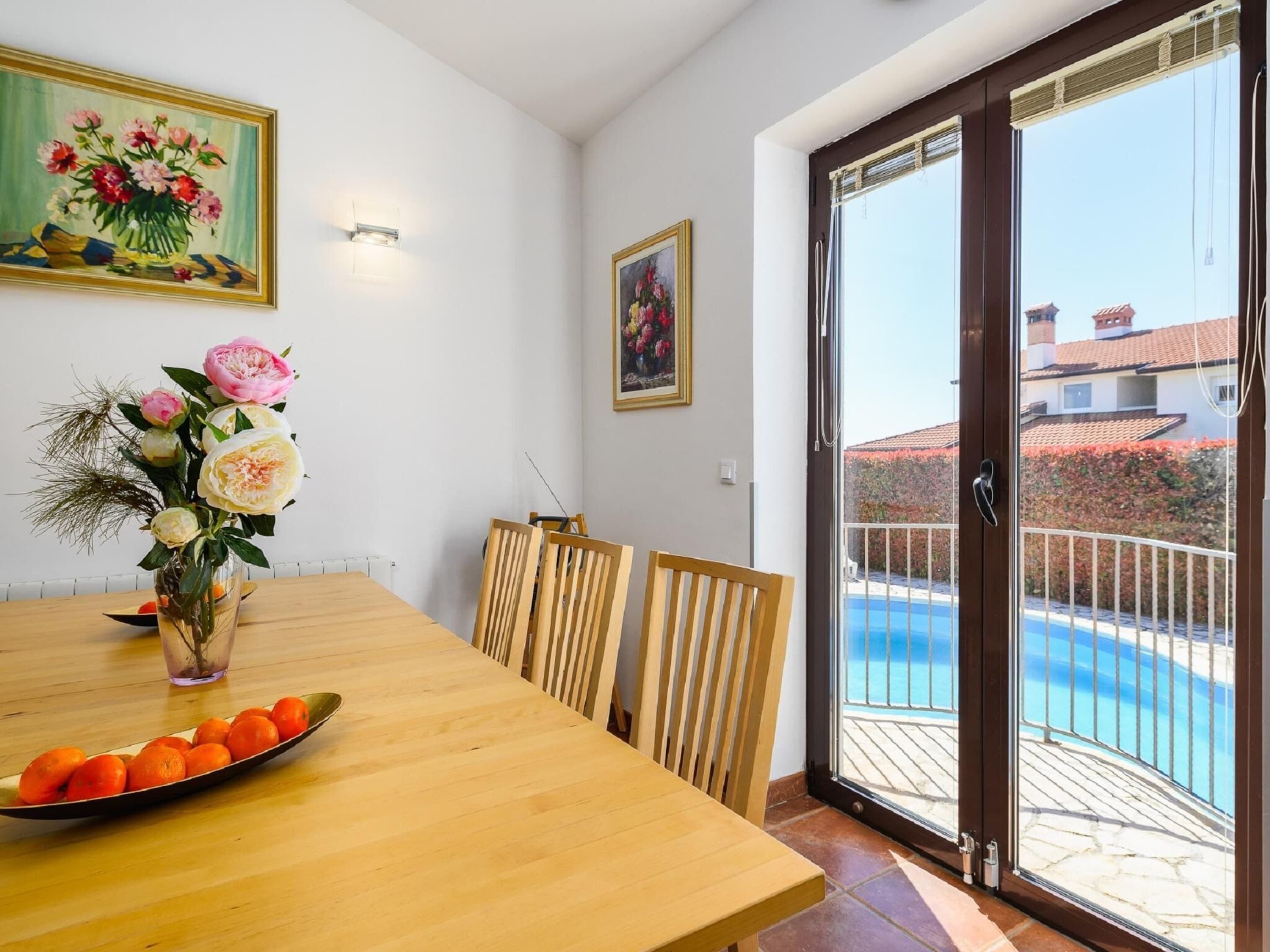 A dining area with a wooden table, chairs, a vase with flowers, and a view of a swimming pool through a glass door.