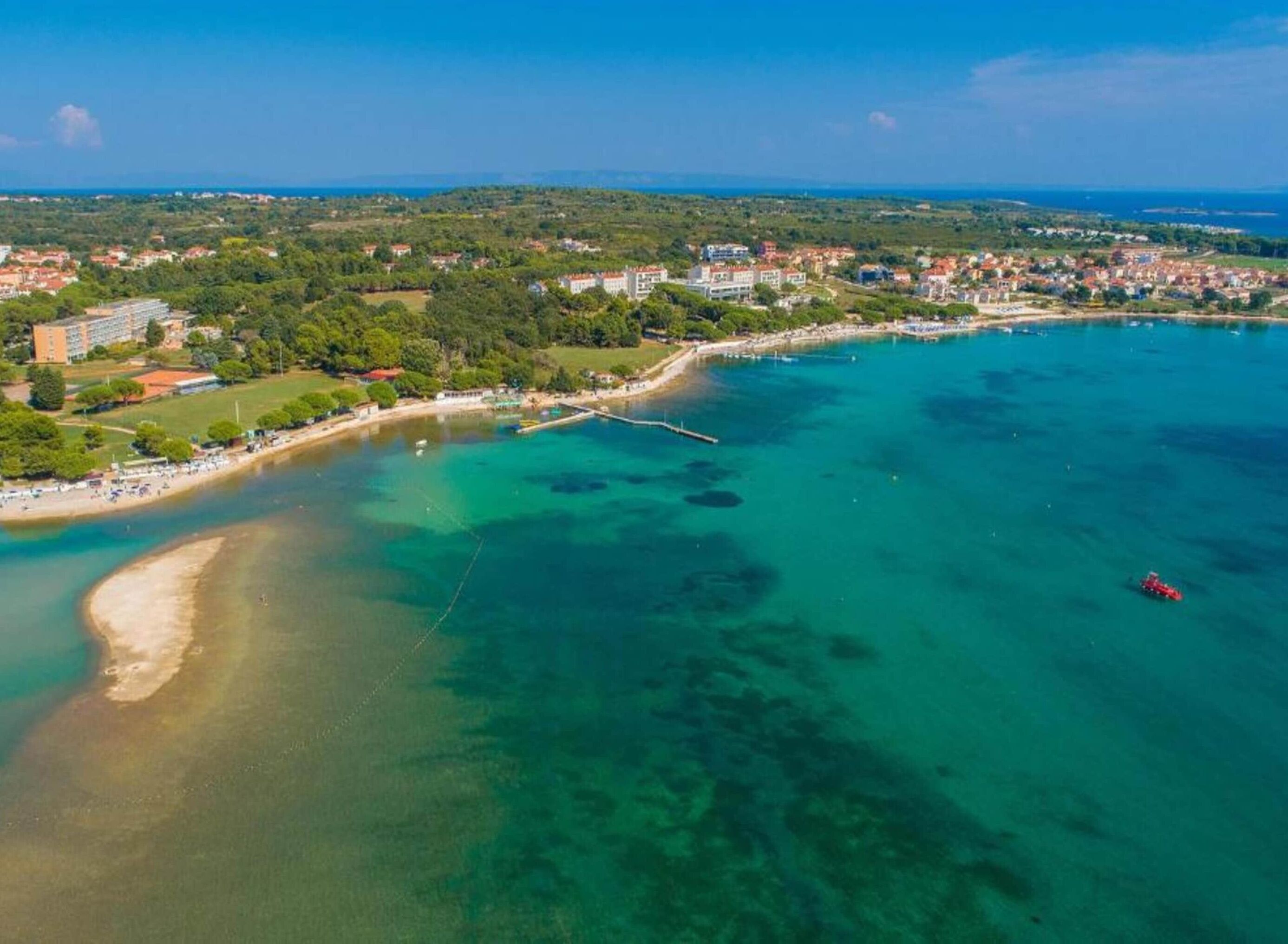 Vue aérienne d’une zone côtière avec une plage, des eaux cristallines et une ville en arrière-plan.