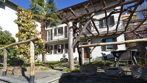 Une cour avec un mur en pierre, une pergola en bois et un bâtiment blanc aux volets bruns.