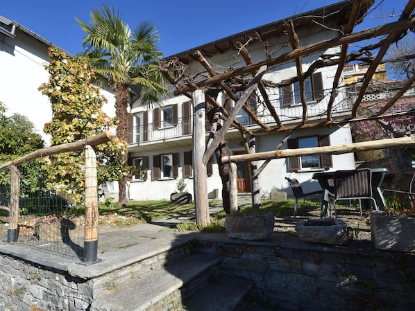 Une cour avec un mur en pierre, une pergola en bois et un bâtiment blanc aux volets bruns.