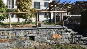 Un mur de pierre surmonté d’une pergola en bois, un arbre en fleurs et un bâtiment blanc avec des volets.