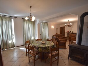 Un coin repas avec une table ronde, des chaises et un lustre. On aperçoit également une cheminée et un canapé au fond.