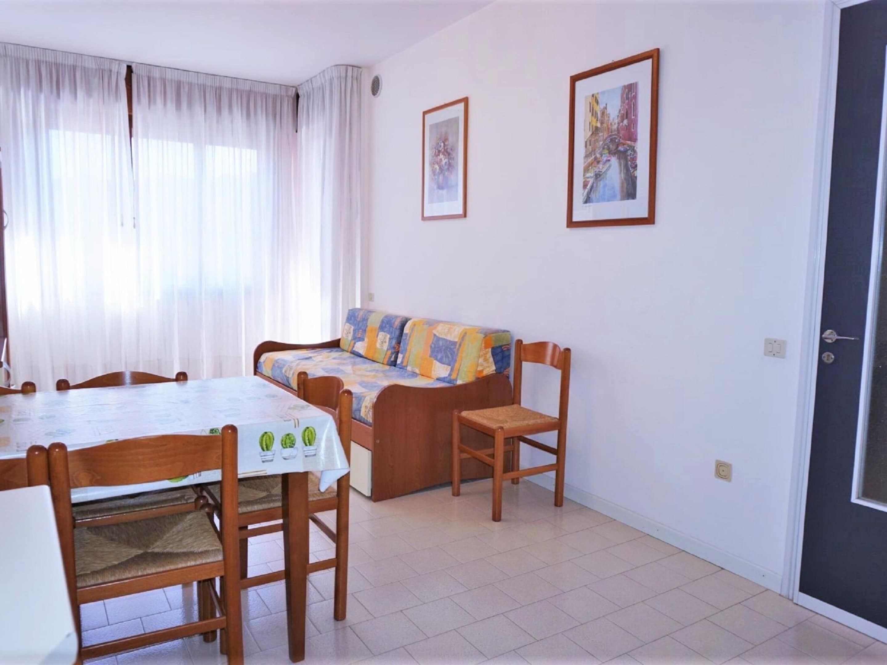 A small, well-lit room with a dining table, chairs, a sofa, and two framed pictures on the wall.