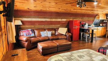 A cozy cabin interior with a wooden ceiling, a leather sofa, a red refrigerator, and a wooden table.