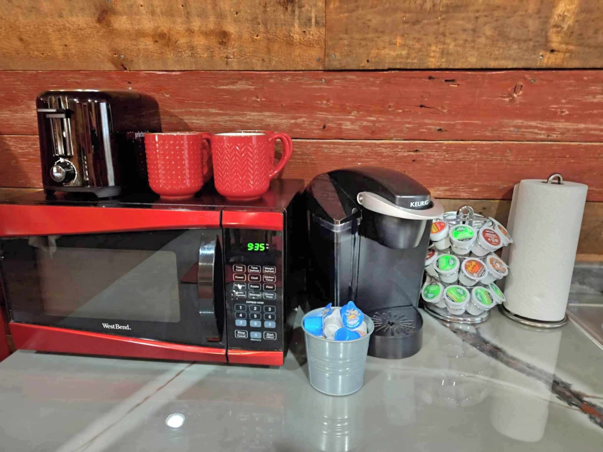 A kitchen with a red microwave, a black coffee maker, two red mugs, a coffee pod holder, and a roll of paper towels.