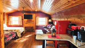 A cozy cabin-style room with a wooden ceiling, a sofa, a small table with stools, a television, and a kitchenette with a refrigerator and coffee maker.