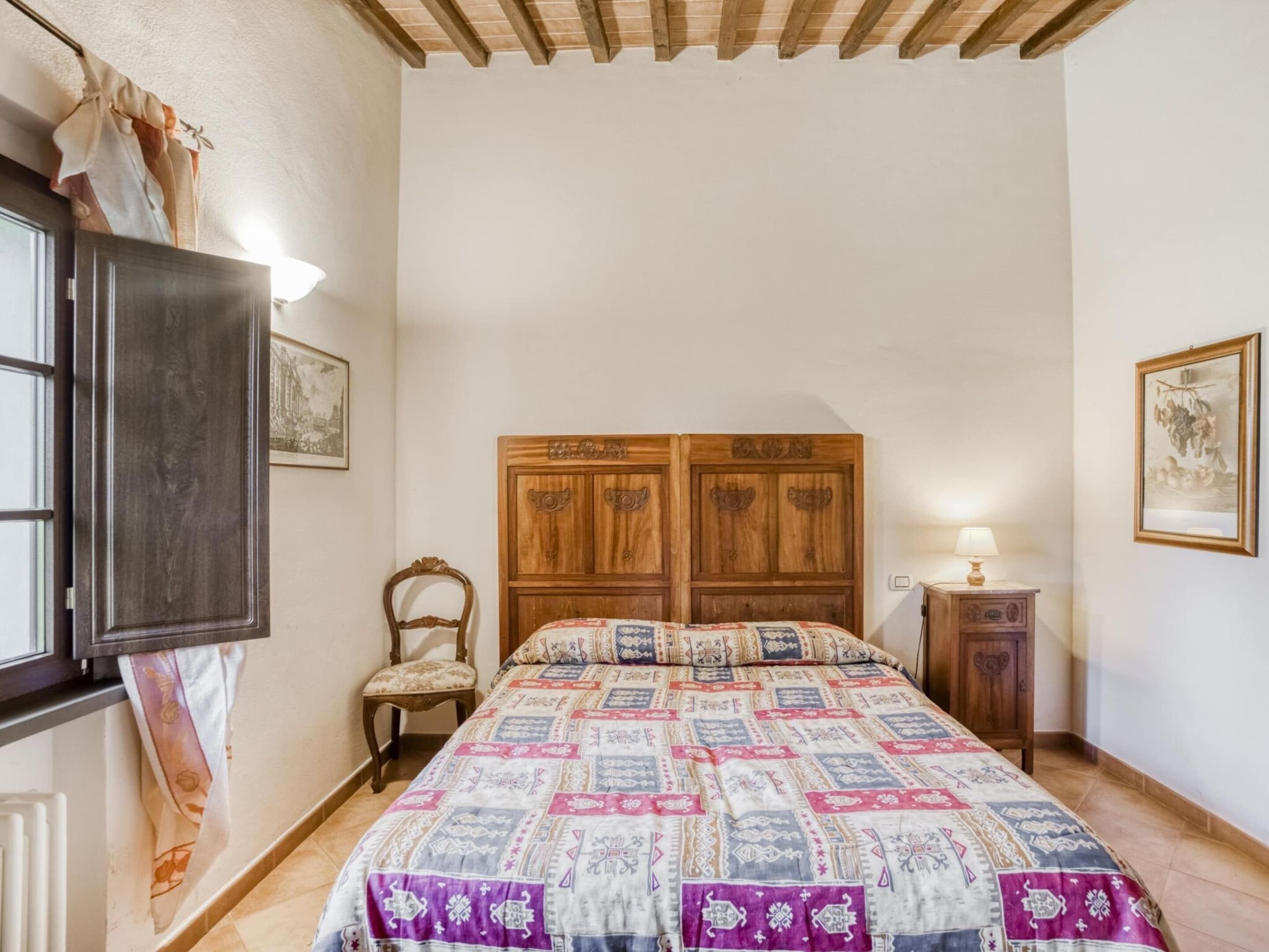 Un dormitorio con una cama doble de madera, una silla, una mesita de noche con lámpara y una ventana con cortinas.