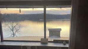 A room with a large window overlooking a body of water, a wooden deck, and a stone fireplace.