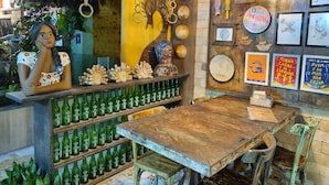Un rústico espacio de bar con estantes de madera llenos de botellas verdes, una mesa de madera y diversos objetos decorativos, incluyendo máscaras y obras de arte enmarcadas.