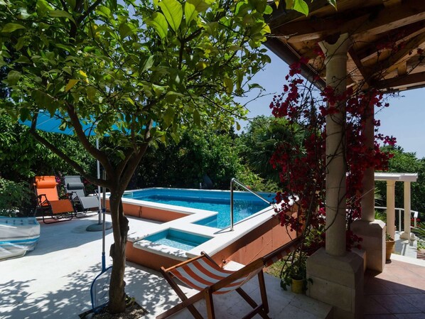 A swimming pool with a small hot tub, surrounded by trees and a pergola with red flowers.