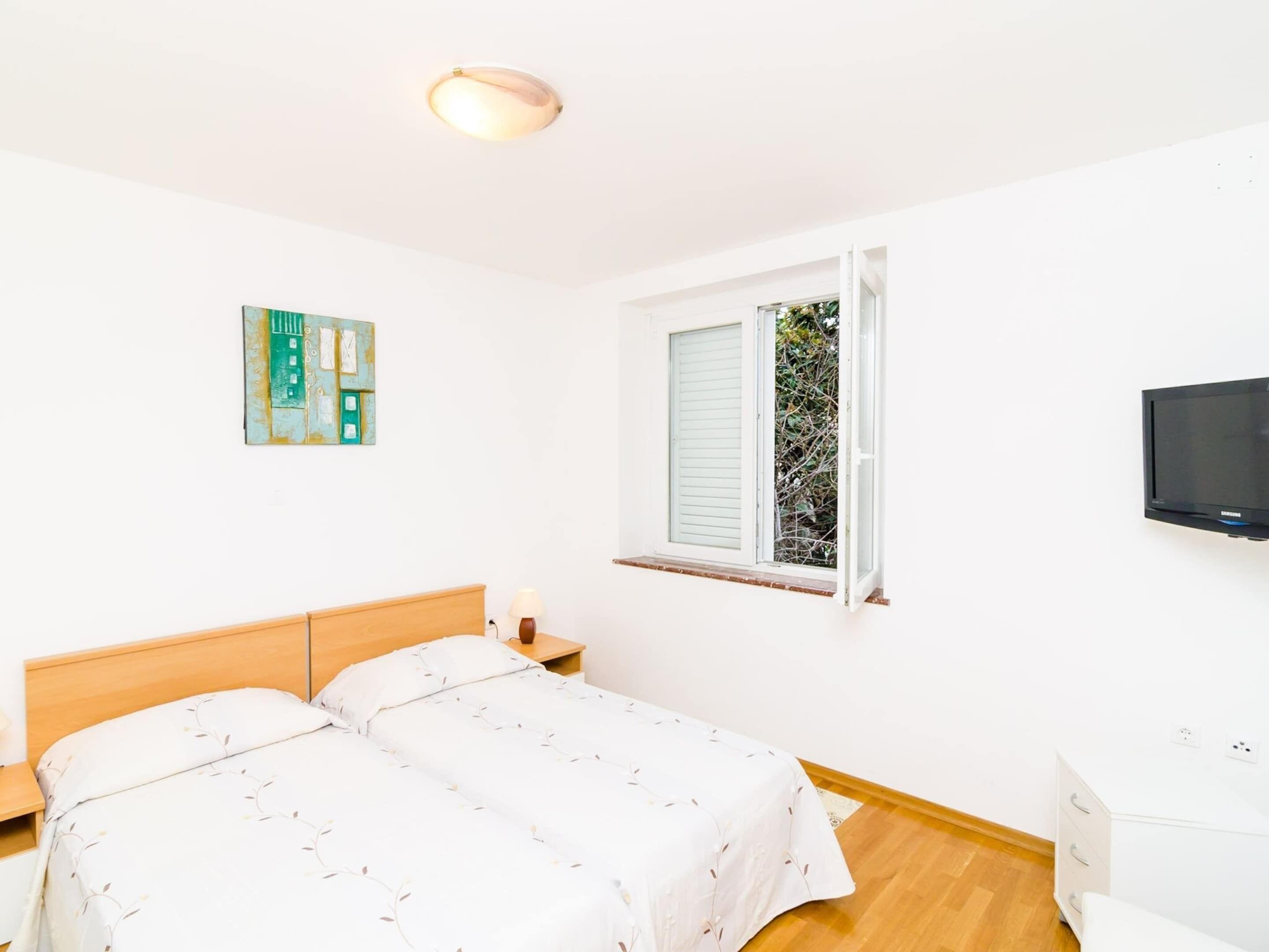 A bedroom with a double bed, a bedside table with a lamp, a window with blinds, a wall-mounted TV, and a small cabinet.