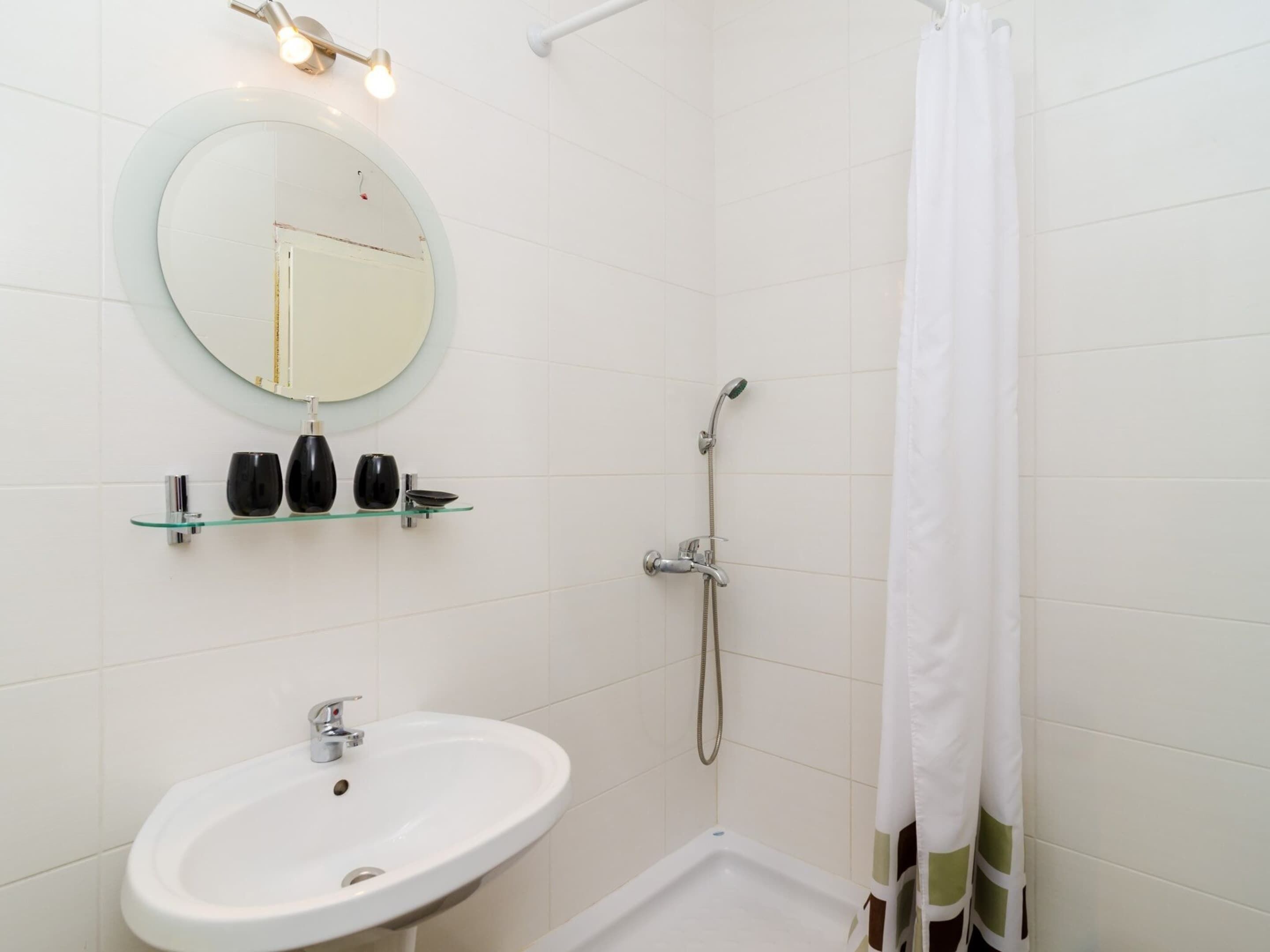 A bathroom with a white sink, a round mirror, a shower with a handheld showerhead, and a glass shelf with toiletries.