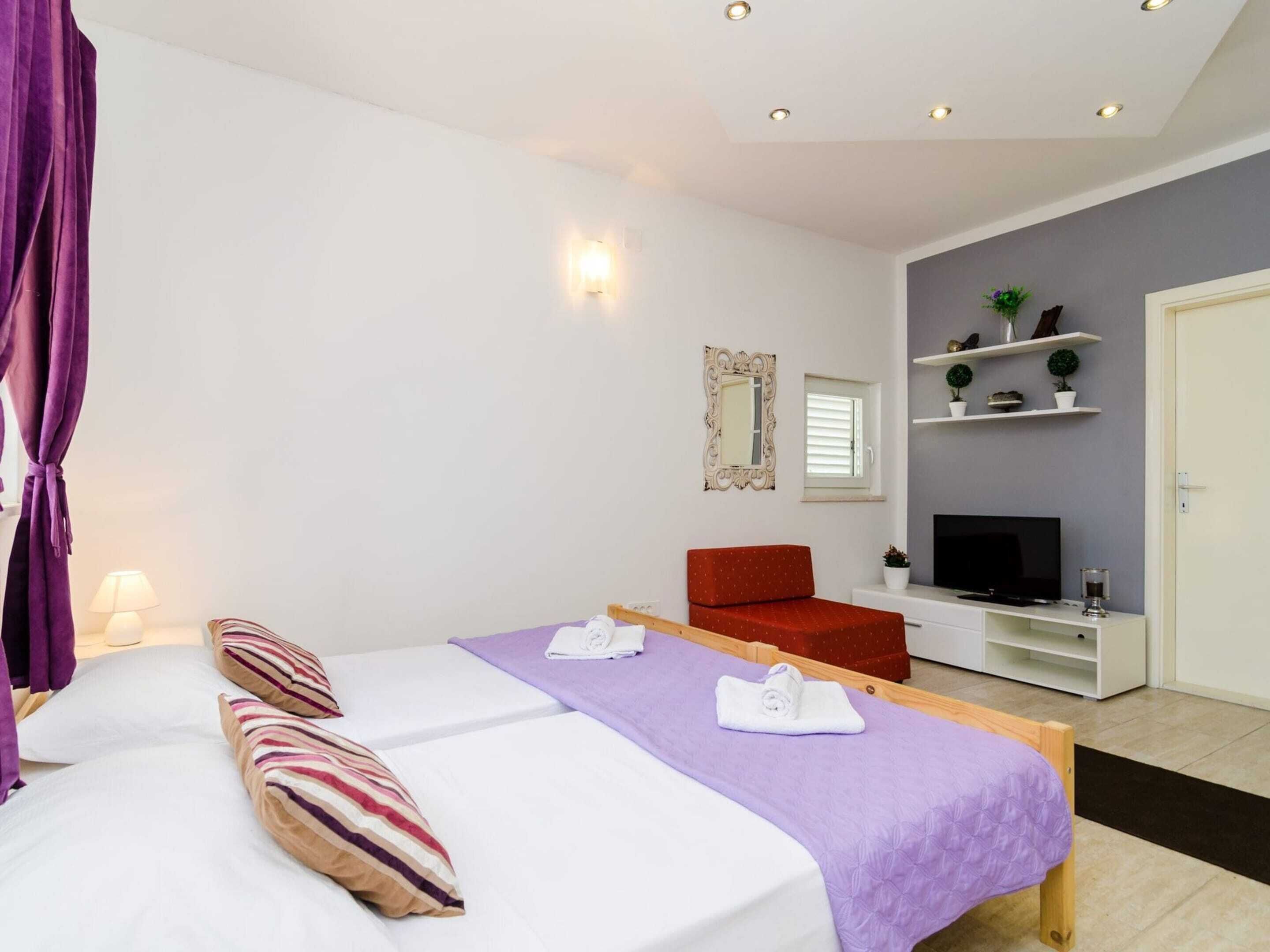 A modern hotel room with a bed, a red chair, a TV, and a shelf with plants.