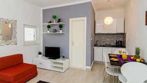A compact living space with a sofa, TV, shelves with plants, a dining area with fruit, and a kitchen.